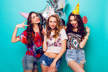 Three beautiful  happy sexy woman  in  stylish summer outfit , paper hats and purity balloons having fun and celebrate  birthday.  Colorful blue background . Pretty girl  hold big lollypop.