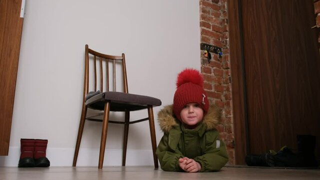 Funny little boy in warm khaki jacket and red knitted hat with pompom lies swinging legs near chair on wooden floor in lobby
