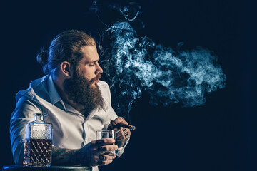 Handsome bearded guy smoke a cigar holds glass of whiskey, blows smoke. Studio shot