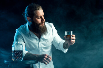 bearded guy holds glass of whiskey, blows smoke on background, dressed in white shirt. Studio shot