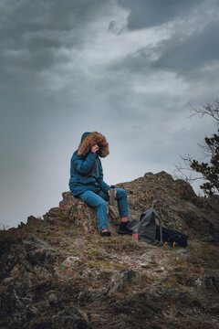 Young woman in a blue down jacket drinks tea from a thermos on a mountain top under a gloomy gray sky