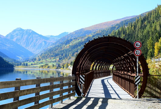 Talumrundung mit Zoggler Stausee und mit Holz verkleideter und wie ein Tunnel &uuml;berdachter Br&uuml;cke, Radweg und Wanderweg im Herbst, Blick vom See &uuml;ber das hintere Ultental mit den Alpen	