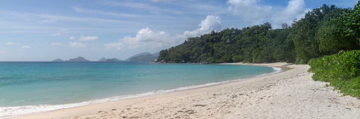 Large-format panoramic photo with high resolution of a typical Seychelles landscape. A perfect snow-white beach. In the distance, on the green hills, there are bungalows with thatched roofs.