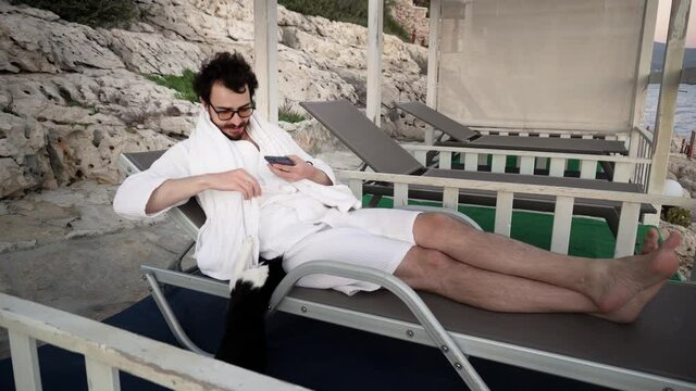 Young caucasian bearded man is resting in lounge area lying on sunbed and playing with a cat trying to take photo of it. A male person is resting on resort under tent on rocky coastline. He is smiling