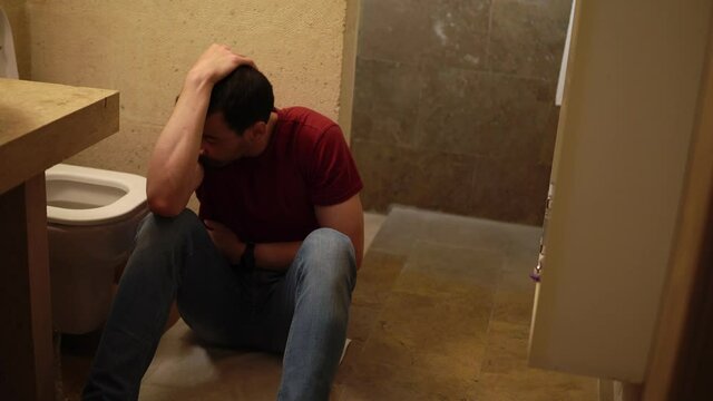 Young sick guy is sitting on the floor of bathroom next to toilet and holding his belly. Caucasian man feels nausea after food poisoning. He is suffering from stomach and head ache and wants to vomit.