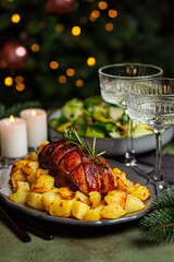 Fototapeta na wymiar Christmas or New Year table. Baked duck roulade stuffed with meat and oranges, baked potato, champagne coupe glasses in front of Christmas tree and burning candles. Salad. Vertical image.