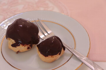 Homemade profiteroles covered with chocolate sauce on the pink tablecloth. Profiterole choux with chocolate ganache on the plate. Delicious eclair dessert with cream and chocolate glaze. 