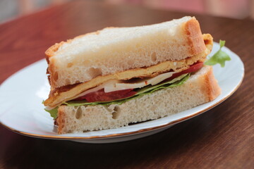 Sandwich with omelette (eggs), lettuce, cheese and tomatoes. Fast, tasty, hearty and nutritious breakfast. Sandwich on the white plate and that plate on the dark wood table.