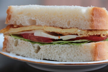 Sandwich with omelette (eggs), lettuce, cheese and tomatoes. Fast, tasty, hearty and nutritious breakfast. Sandwich on the white plate and that plate on the dark wood table.