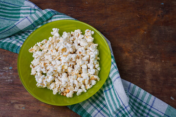 Green plate of salted popcorn on wooden table background with kitchen towel. Top view, copy space