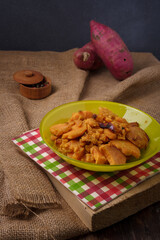 Green plate on burlap with stewed sweet potatoes or yam. Tasty dish, healthy country food. Pepper peas spice pot, raw potatoes on background