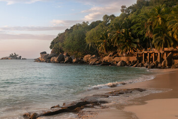 Evening photo of a deserted Seychelles beach. Perfectly smooth sand is washed by the sea surf. The beach ends with a granite cliff and palm trees growing above it. Tropical forest in twilight shadows