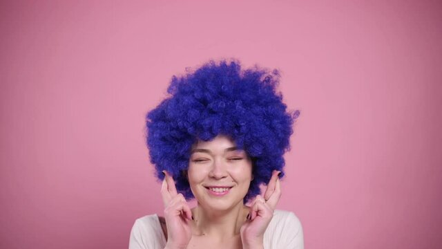 Funny young woman with blue hair makes wish with her fingers crossed. Portrait on pink background.