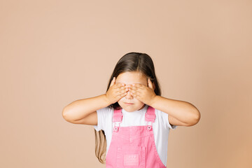 Close up portrait rebellious kid closing eyes with hands not see others with closed mouth not talk with others wearing bright, pink jumpsuit and white t-shirt on beige background.