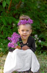 Full length body size smiling cute caucasian little girl child kid of 5 years with lilac wreath on head and lilac bouquet in hands looking at camera squatting in green park on trees background