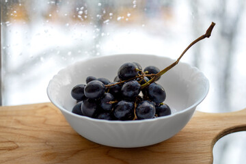 Black and blue grapes in drops of water in a white plate against the background of rain