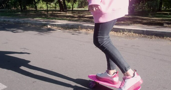A young girl in a pink sweater tries to ride a skateboard on the road in the park, constantly jumps from a skid to the side.