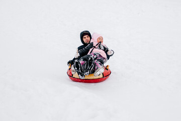 Fototapeta na wymiar kid smiling and sliding down hill on sled with father looking away wearing warm winter clothes in forest. Astonishing background full of white color and snow. 