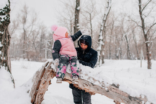 Distant photo of female kid sitting on tree log wearing pink winter clothes throwing snow at father standing behind daughter in forest. Astonishing background full of white color and snow. 