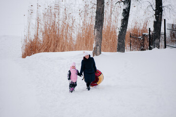 Fototapeta na wymiar Distant photo of female kid and mother with sled both looking into camera and walking towards camera in snow in forest. Astonishing background full of white color and snow. 