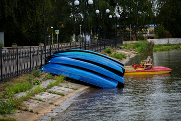 blue overturned boats on the pier on a cloudy summer day, against a background of wrought-iron hedges and green trees. Catamaran and boats.
