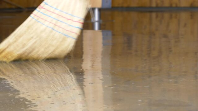 LOW ANGLE, CLOSE UP: Person uses a straw broom to sweep the flooded floor after an episode of heavy rain. Broom with rough straw bristles sweeps away the dirty water covering the basement floor.