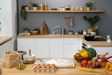 Fototapeta na wymiar Interior of kitchen with wooden table, fresh vegetables, sifted flour, eggs, bottle of olive-oil and bunch of spaghetti