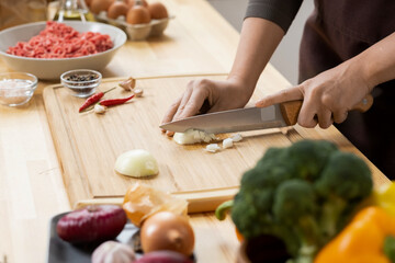 Hands of young woman cutting raw onion with sharp kitchen knife on wooden board while cooking food