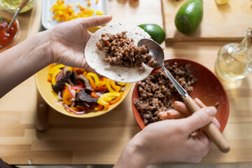 Hands of young woman with spoon putting fried minced meat on taco tortillas while cooking national food