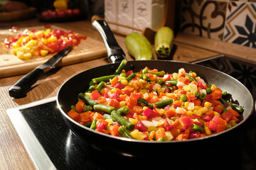 Mixture of fresh chopped vegetables on frying-pan standing on electric stove by kitchen table with knife and wooden board