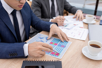 Hand of young Asian businessman in formalwear pointing at financial data while looking through papers by table