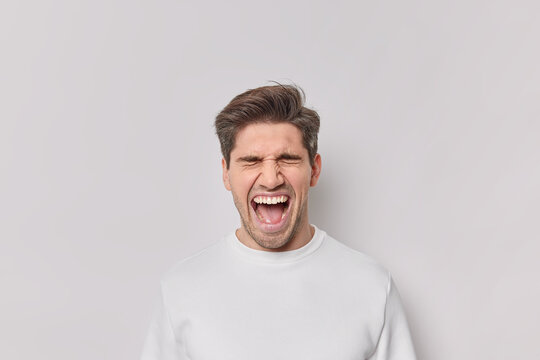 Portrait of emotional man screams from amazement keeps mouth wide opened yells loudly wears casual jumper isolated over white background. Excited male model reacts on awesome news or surprise