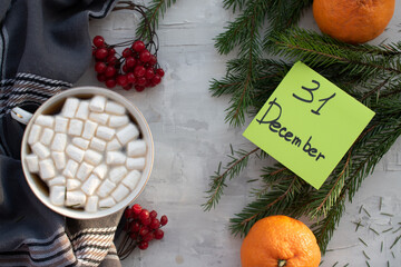 On a gray background, a gray plaid, a cup of cocoa and marshmallows, twigs of spruce, sprigs of viburnum, tangerines and the inscription December 31. New Year and Christmas background.