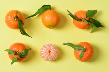 Six mandarins lie next to each other against a yellow background. There are still some leaves on the mandarins. A tangerine is peeled.