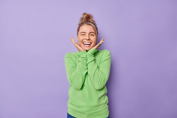 Positive young blonde European woman winks eye and smiles broadly keeps hands under chin feels...