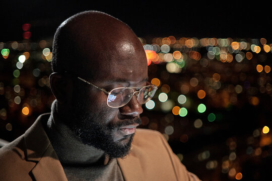 Young bald businessman in eyeglasses and formalwear standing in front of camera against night traffic lights