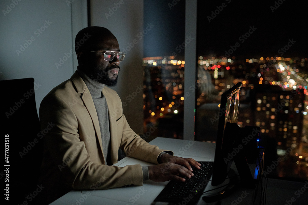 Poster Bearded African office worker in formalwear typing and looking at computer screen while working overtime at night