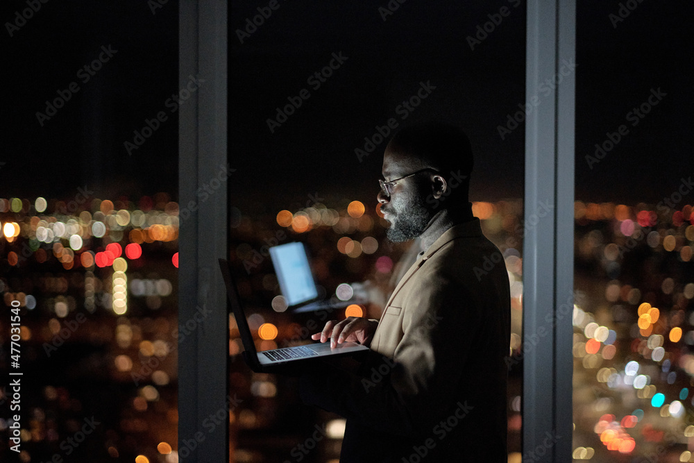 Poster young businessman with laptop standing against large window in office and analyzing online data late