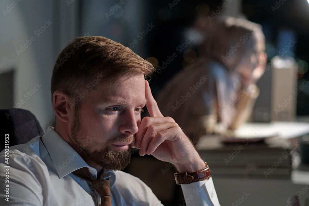 Poster serious young businessman in formalwear sitting in front of computer and looking through online data