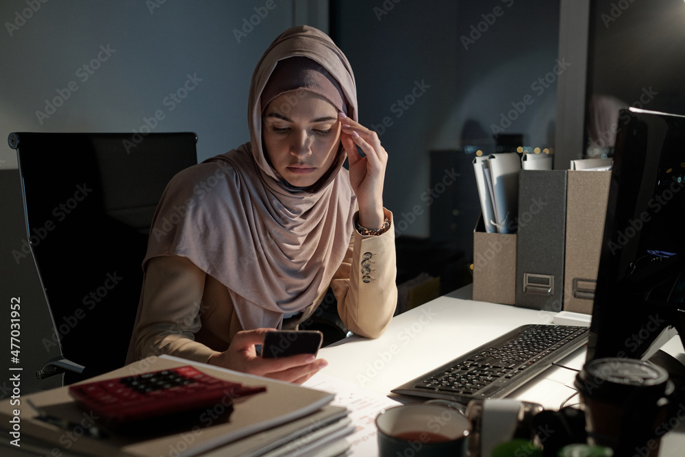 Aufkleber young serious businesswoman in hijab scrolling in smartphone late at night while sitting by desk in 