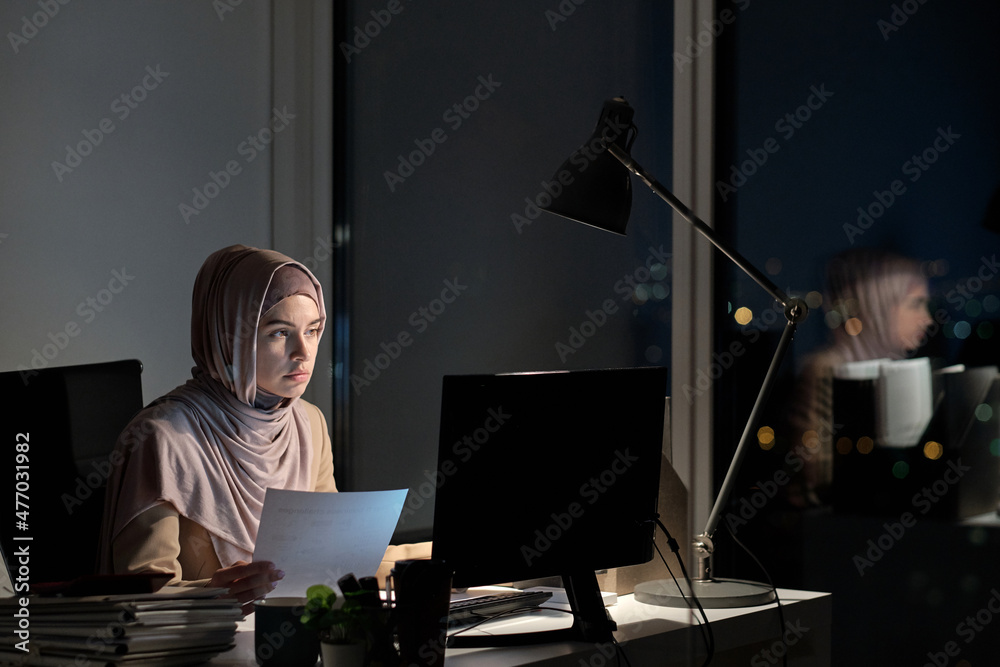 Aufkleber young muslim female in hijab holding paper and looking at computer screen while sitting by desk and 