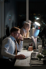 Young elegant male economist looking at computer screen while sitting by desk in office against Muslim female colleague