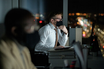 Young contemporary male accountant in protective mask looking at computer screen while working late at night