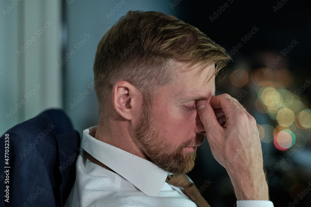 Poster side view of face of young tired businessman sitting by workplace in office and working overtime at 