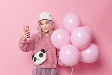 People and holidays concept. Charming Asian woman takes selfie via smartphone poses with inflated balloons has fun on party makes memorable photo wears outerwear isolated over pink background