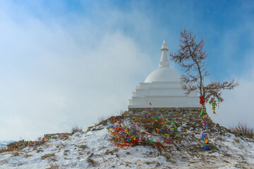 White Buddhist stupa and ritual trees with colorful ribbons on a cloudy winter day at the top of the sacred island of Ogoy. Lake Baikal, Siberia, Russia