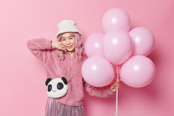 Pleased lovely Asian girl with pink hair wears white panama and fur coat holds bunch of inflated helium balloons celebrates something poses against rosy background. People and holidays concept
