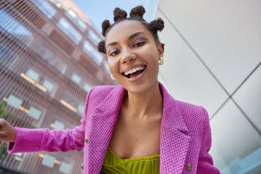 View from below of cheerful fashionable woman smiles broadly enjoys life poses in urban setting during daytime has bun hairstyle wears stylish clothes feels satisfied. People lifestyle concept