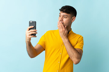 Young Colombian man using mobile phone isolated om blue background with surprise and shocked facial...