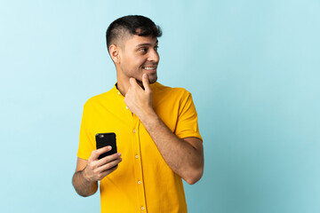 Young Colombian man using mobile phone isolated om blue background thinking an idea and looking side